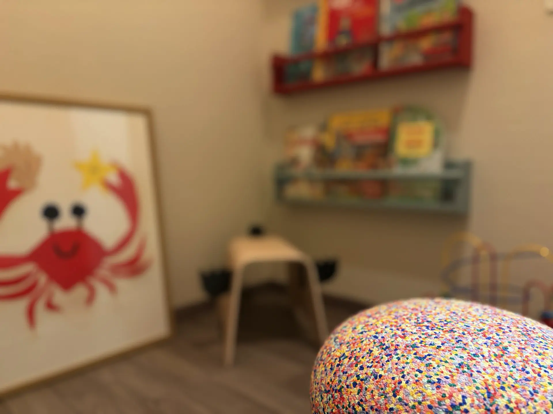 Nahaufnahme eines bunten, gepunkteten Sitzpolsters in einem Kinderzimmer mit einem Spielstuhl, Bücherregalen und einem Bild einer roten Krabbe an der Wand.