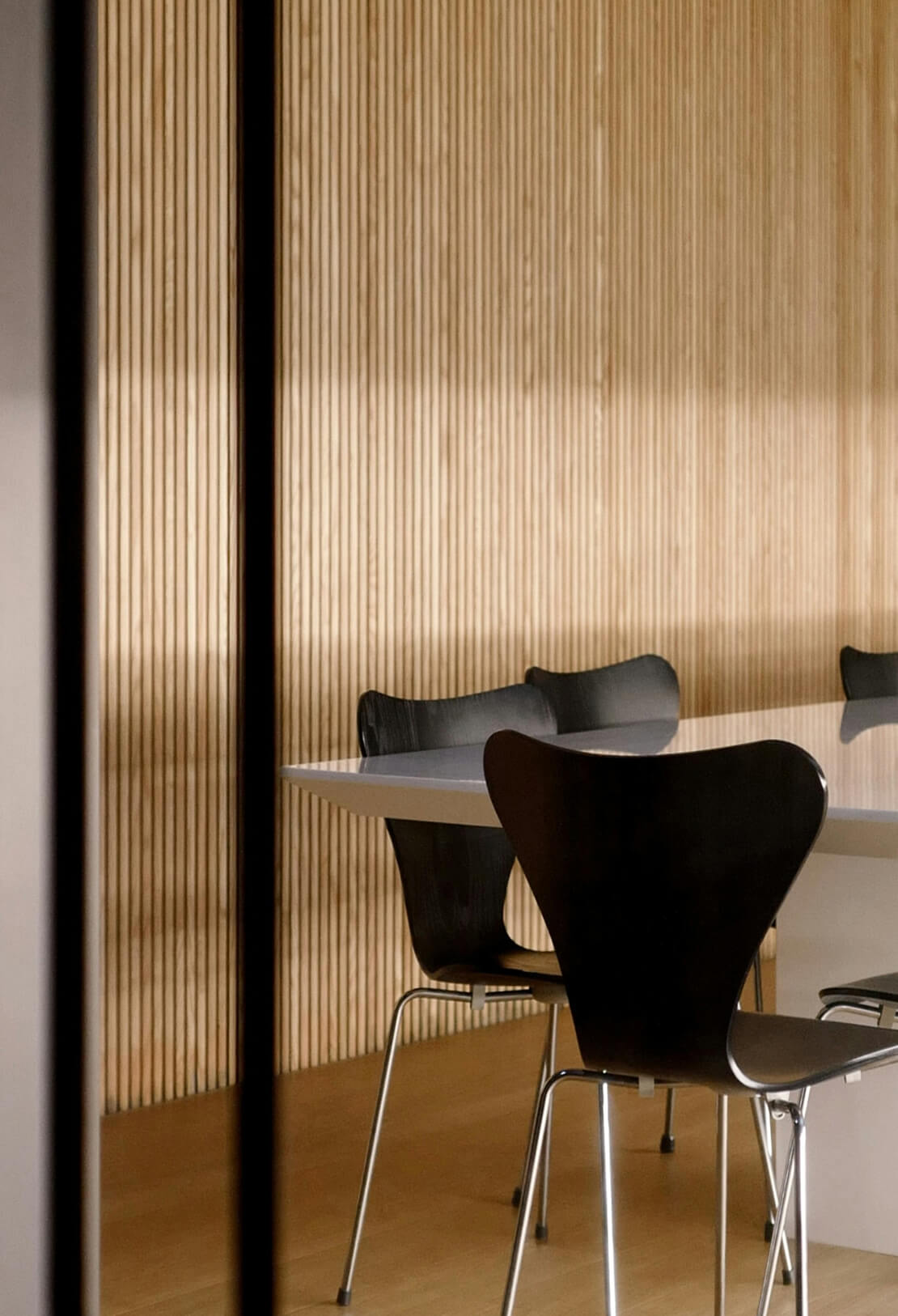 Minimalist interior featuring a light wooden wall with vertical slats, a white table, and stylish black chairs.
