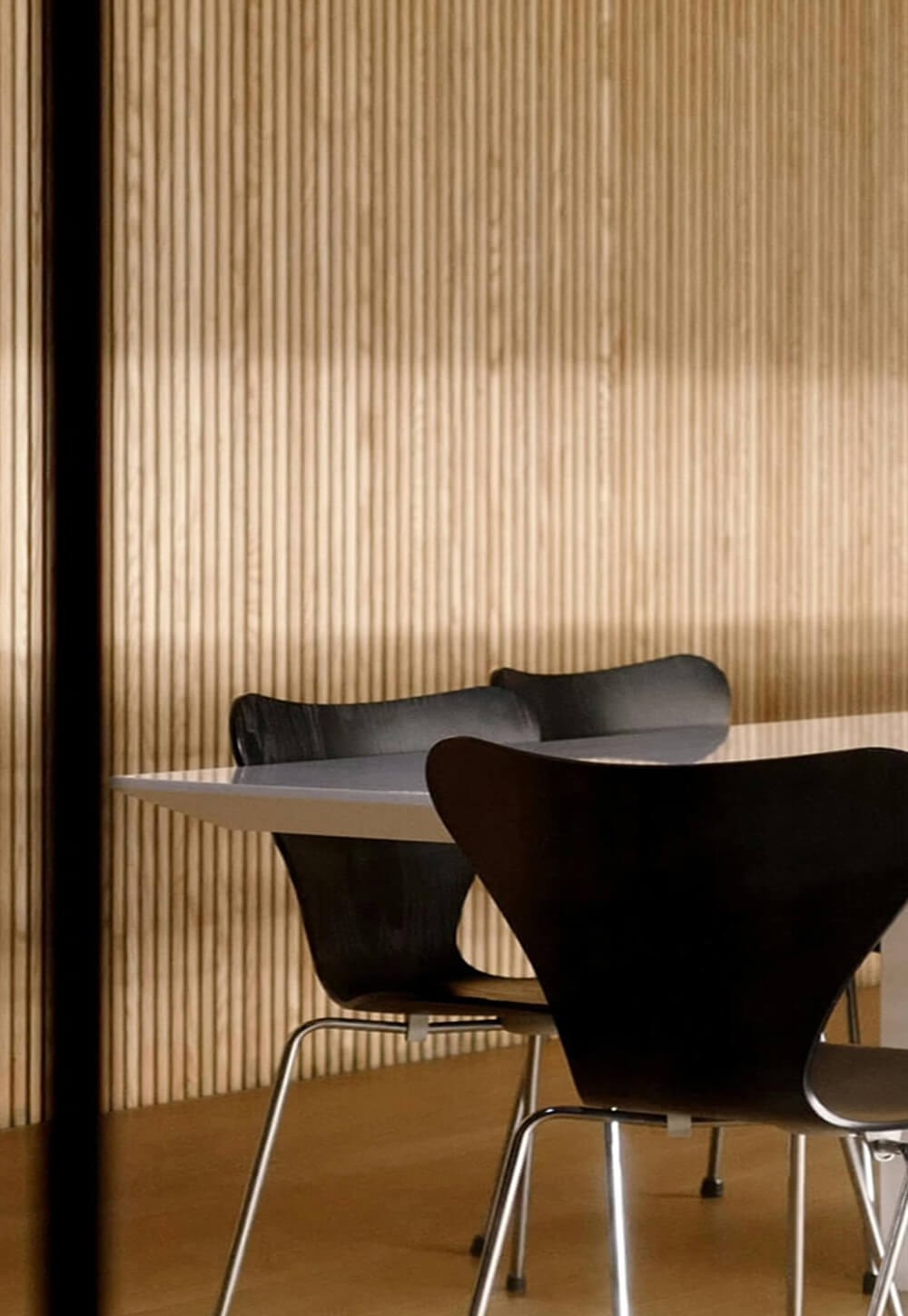 Modern interior scene featuring a minimalist table and black chairs against a textured wooden wall. Soft natural lighting enhances the design.