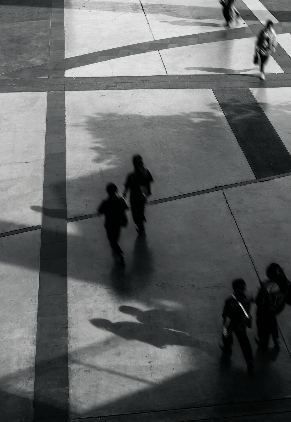 Black and white image of people walking on a geometric patterned floor, casting shadows in various directions.