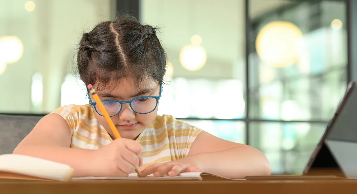 Young girl with braided hair and glasses focused on writing in a notebook at a desk.