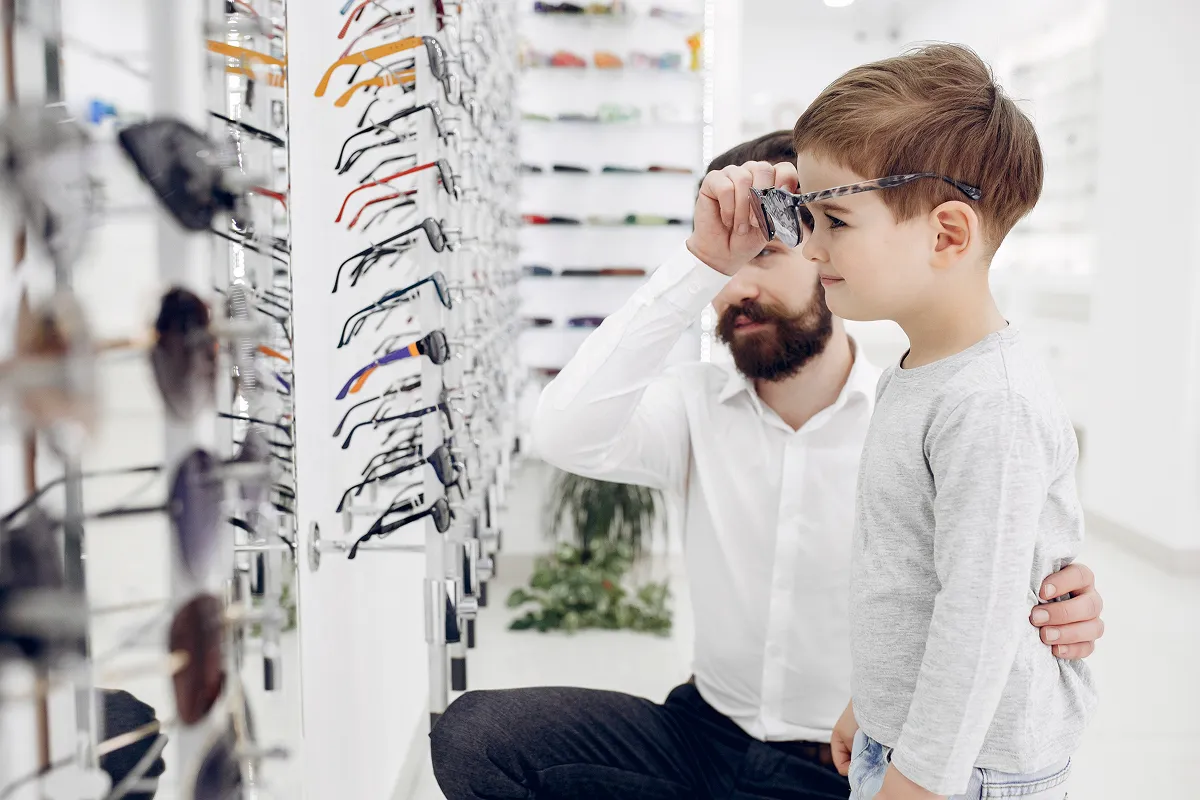 Man helping young boy try on eyeglasses in an optical store with shelves of glasses in the background.
