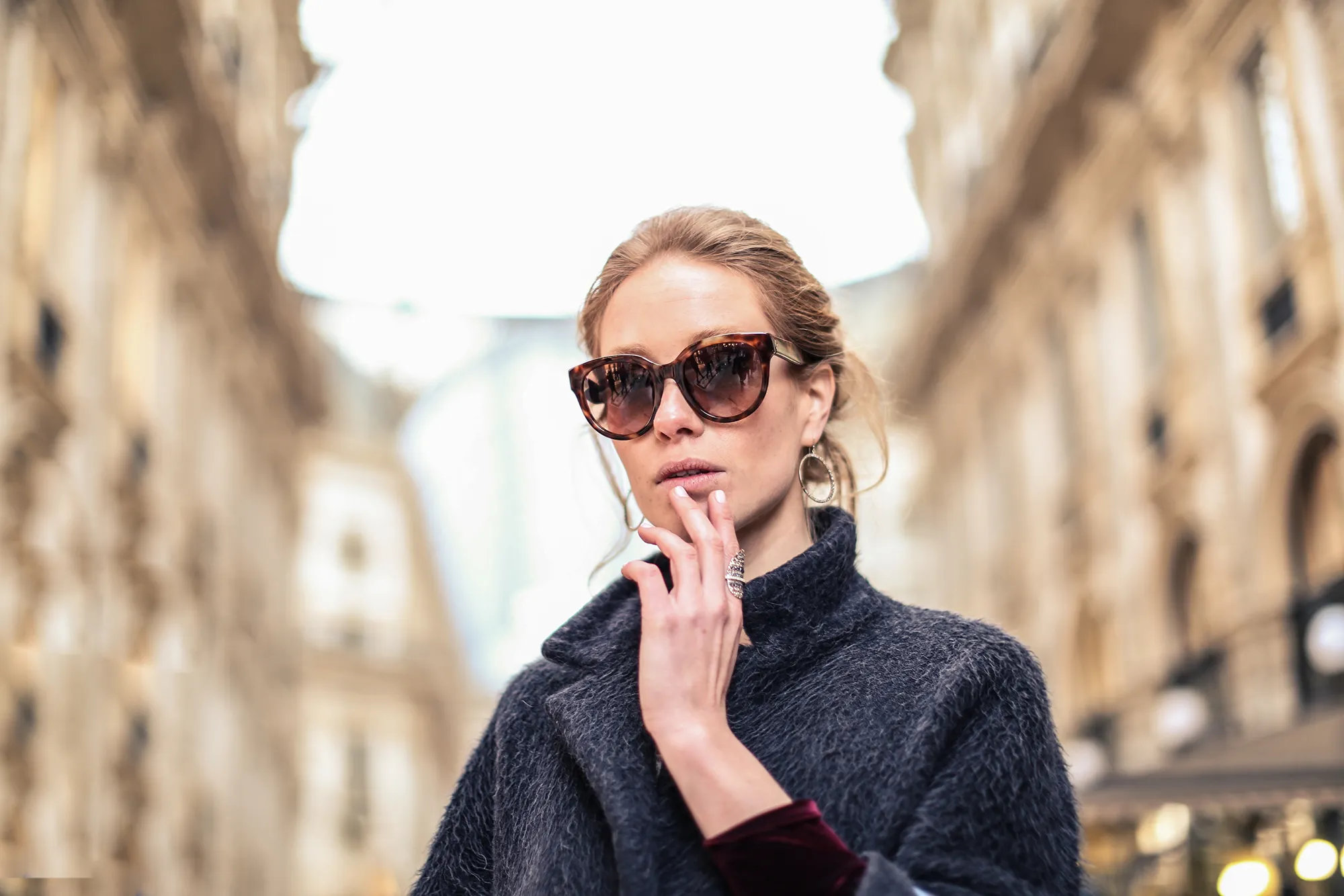 Woman wearing large tortoiseshell sunglasses and a dark wool coat standing outdoors on a blurred city street.