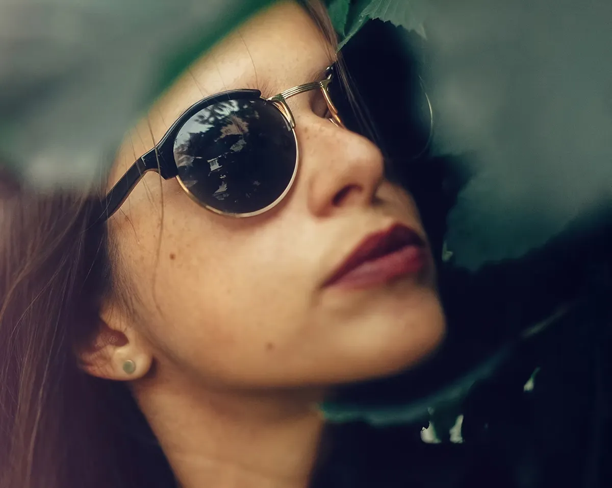 Close-up of a woman wearing round sunglasses partially covered by green leaves.