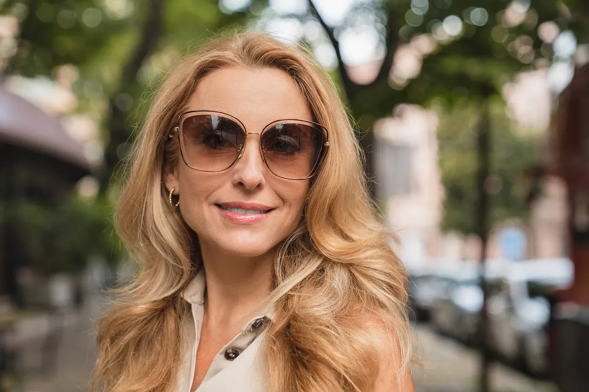 Smiling middle-aged woman with long blonde hair wearing large sunglasses and beige sleeveless top outdoors.
