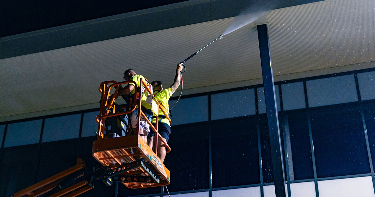 Person from Hosed Building and Gutter team, power washing a large sidewalk area near buildings with Vans and MG Motor signage in the background.