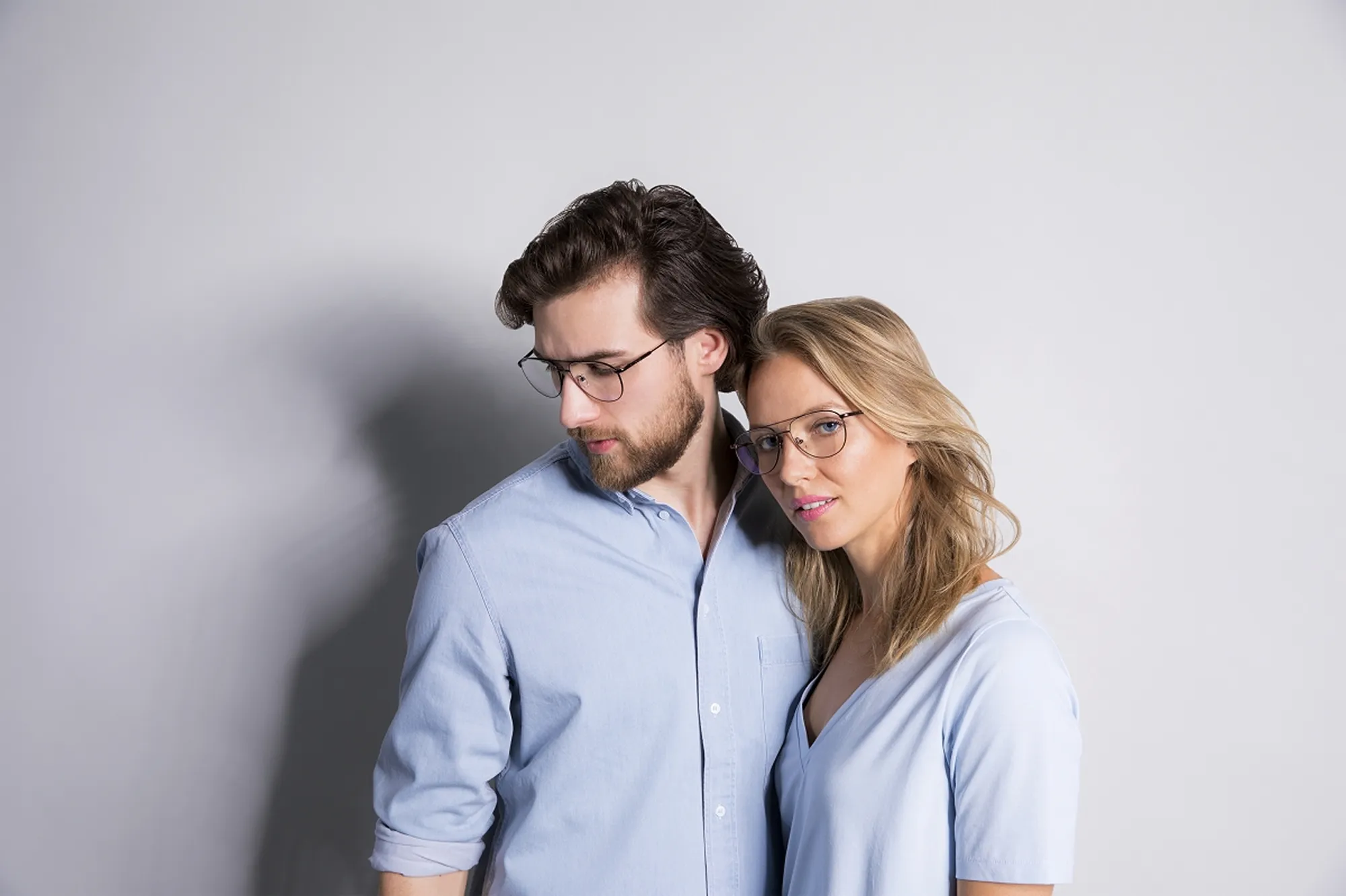 Man and woman wearing eyeglasses and light blue shirts standing close together against a plain gray background.