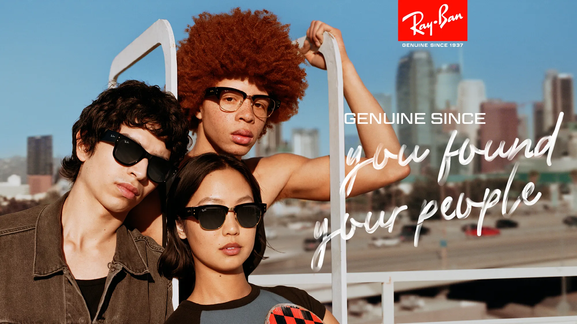 Three young adults wearing Ray-Ban sunglasses posing outdoors with a city skyline in the background.