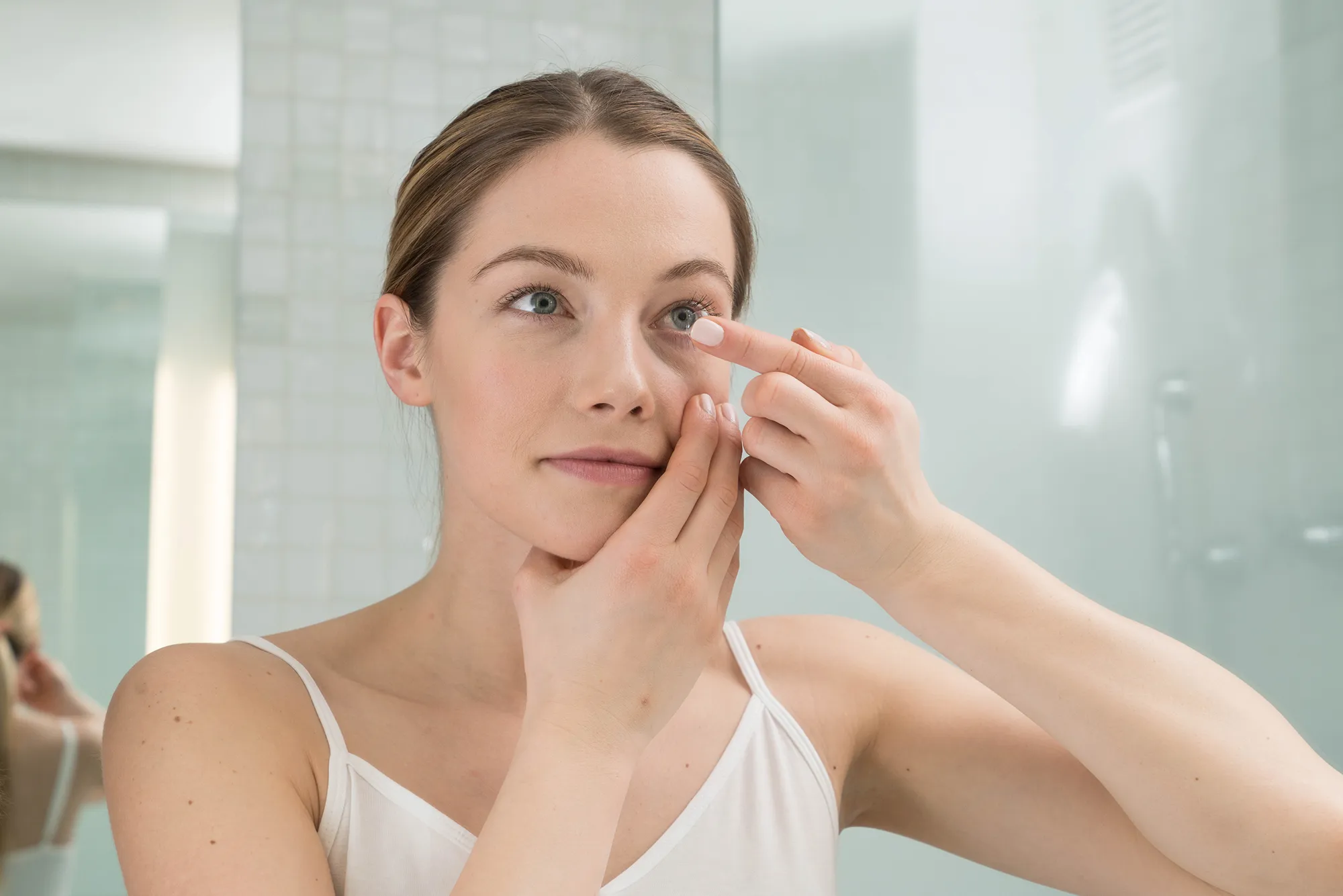 Woman in a white tank top putting in a contact lens in her right eye in a bathroom with tiled walls.