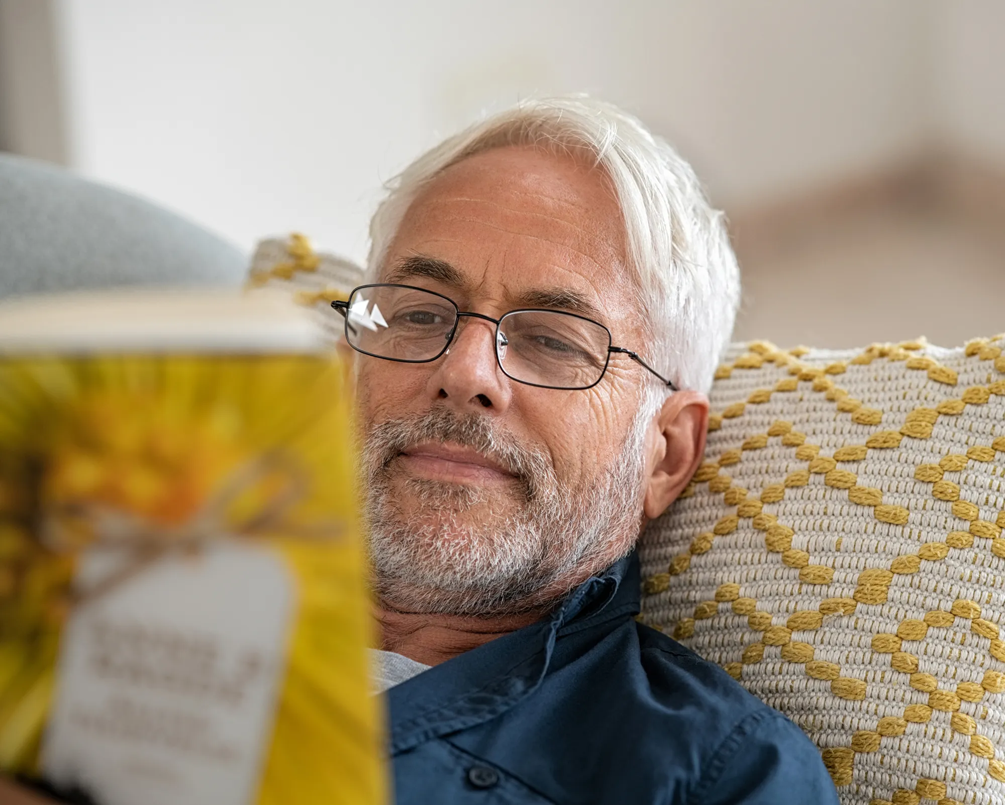 Smiling older man with glasses reclining on a patterned cushion while reading a book.