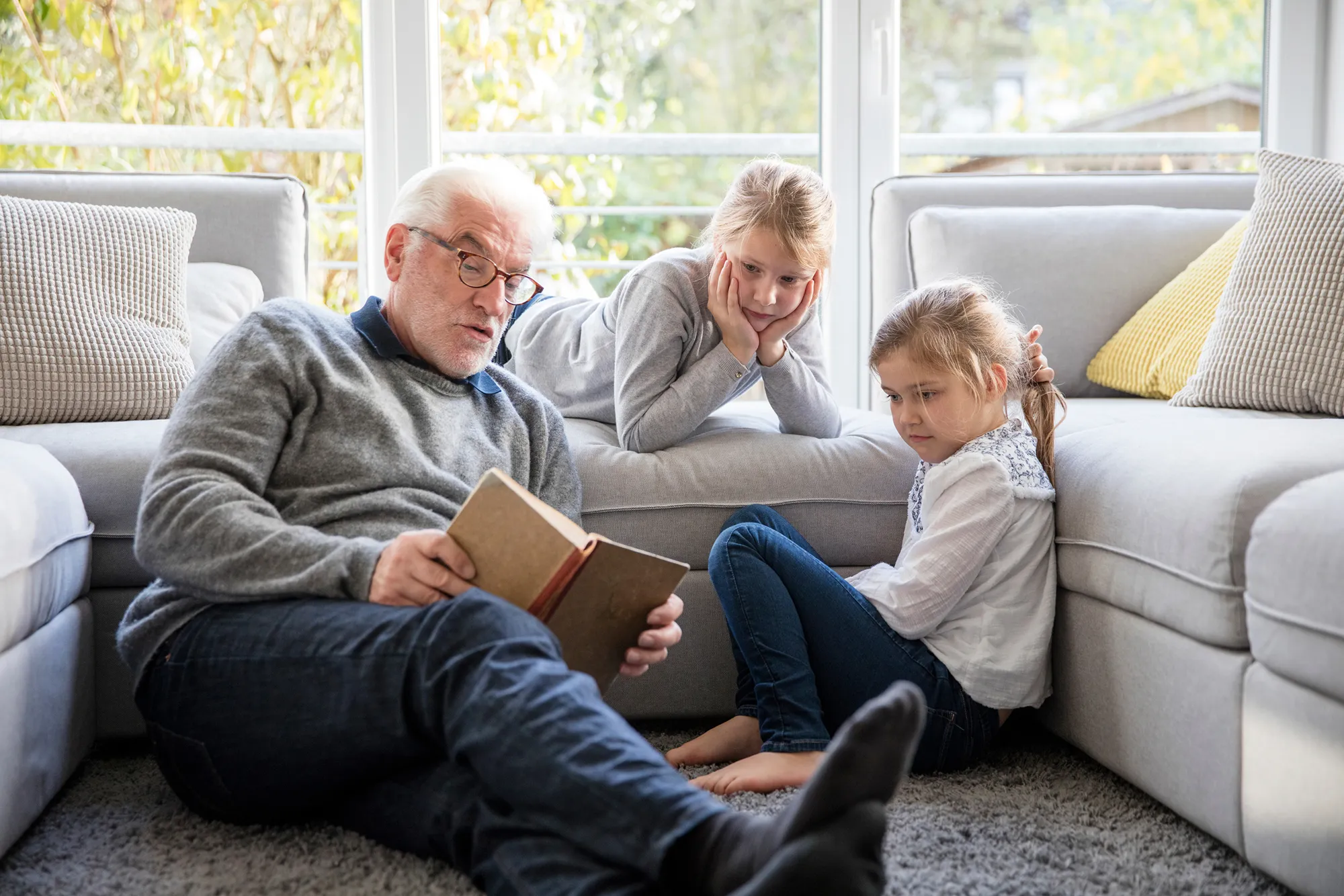 Elderly man reading a book to two young girls sitting on a carpet in a bright living room.