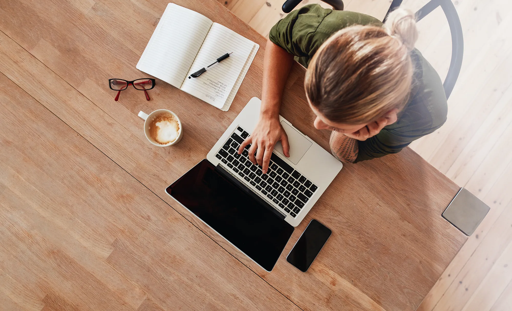 Woman working on a laptop at a wooden table with coffee, glasses, and a notebook nearby.