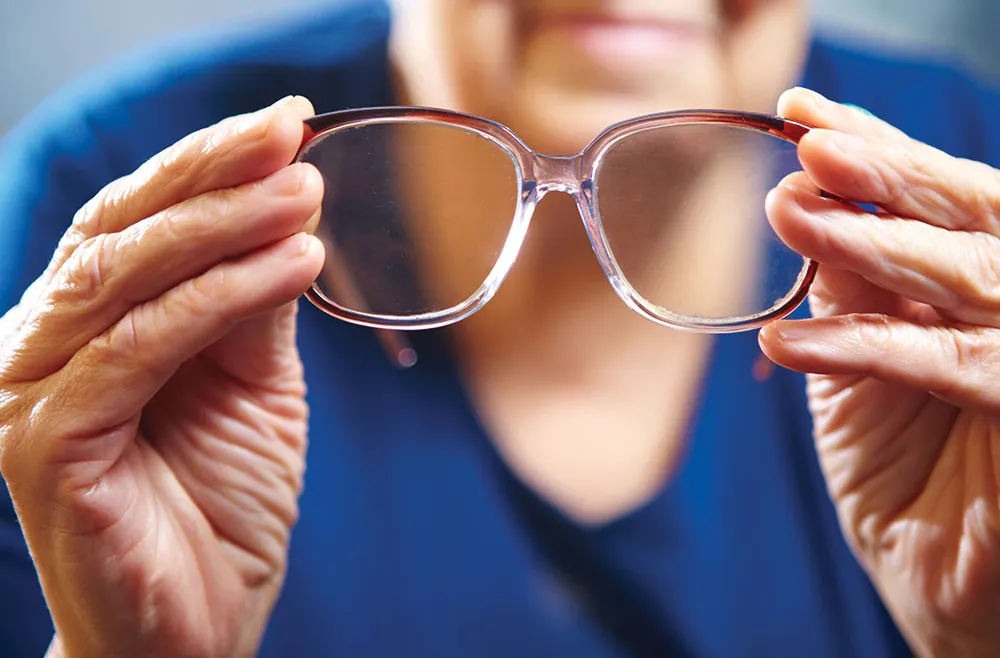 Elderly person holding a pair of eyeglasses with both hands in front of a blurred face.