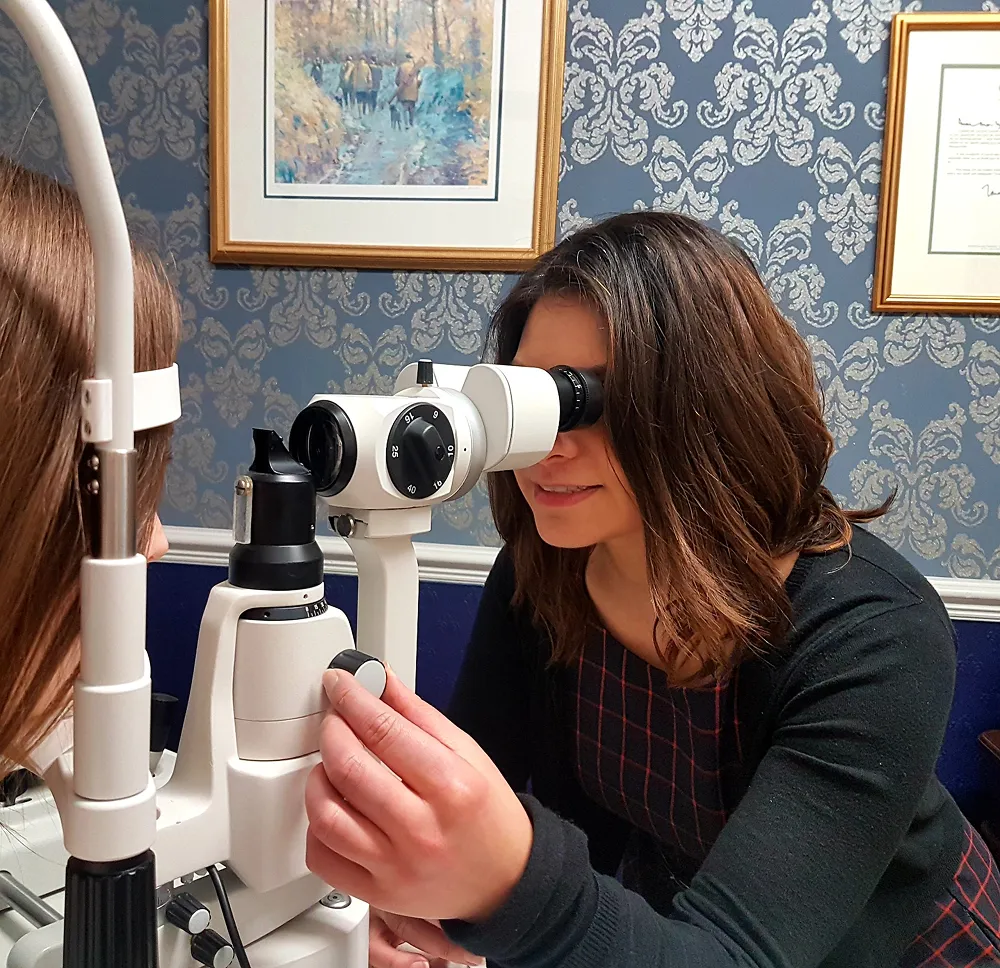 Optometrist using a slit lamp to examine a woman's eyes in a decorated office room.