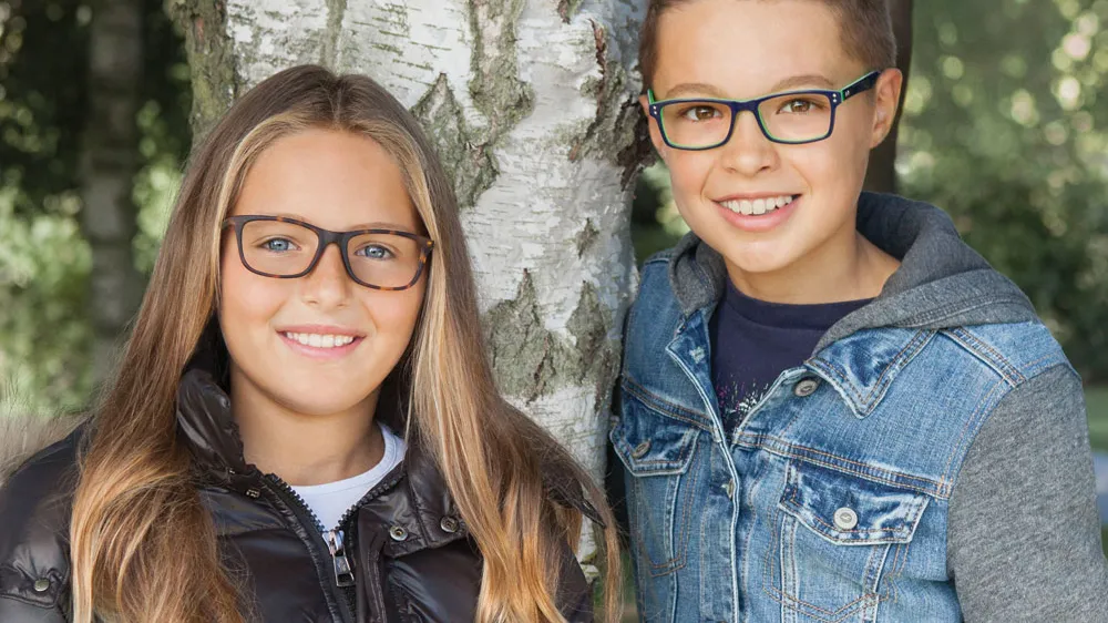 Two smiling children wearing glasses outdoors in front of a tree.