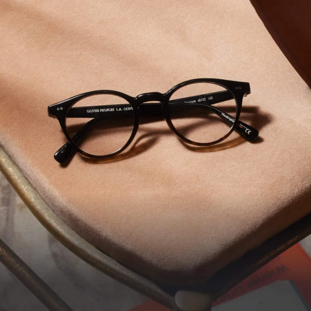 Round black Oliver Peoples glasses frames resting on a tan suede chair seat.