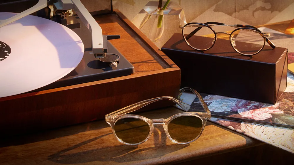 Two pairs of glasses — clear-framed sunglasses and round gold-rimmed frames — styled on a wooden surface beside a record player.