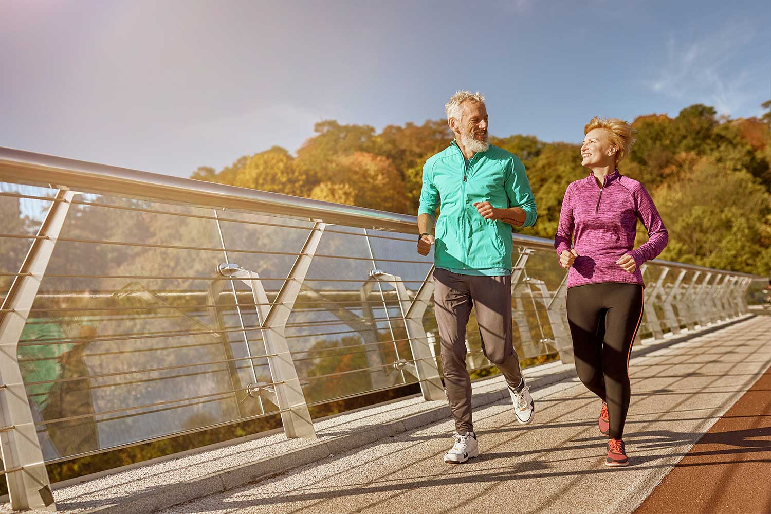 Smiling elderly couple jogging together on a sunny outdoor pathway with autumn trees in the background.