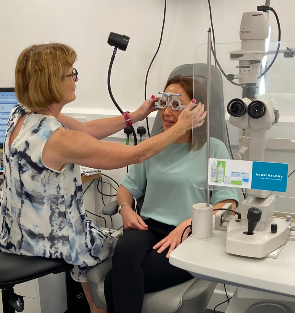 Optometrist fitting trial frame on patient in eye exam room with eye testing equipment nearby.