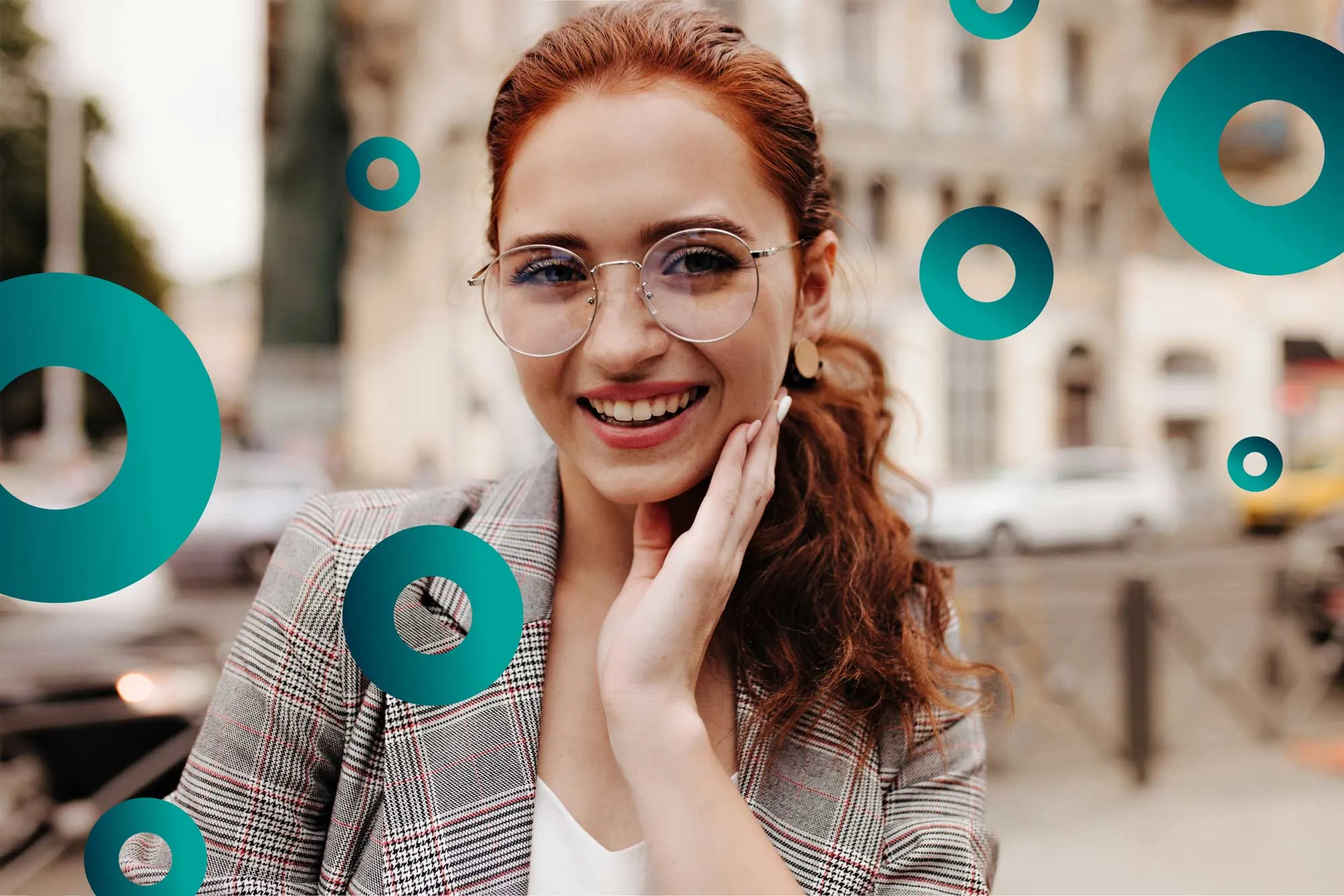 Smiling red-haired woman with glasses and checkered blazer touches her cheek outdoors in a city.