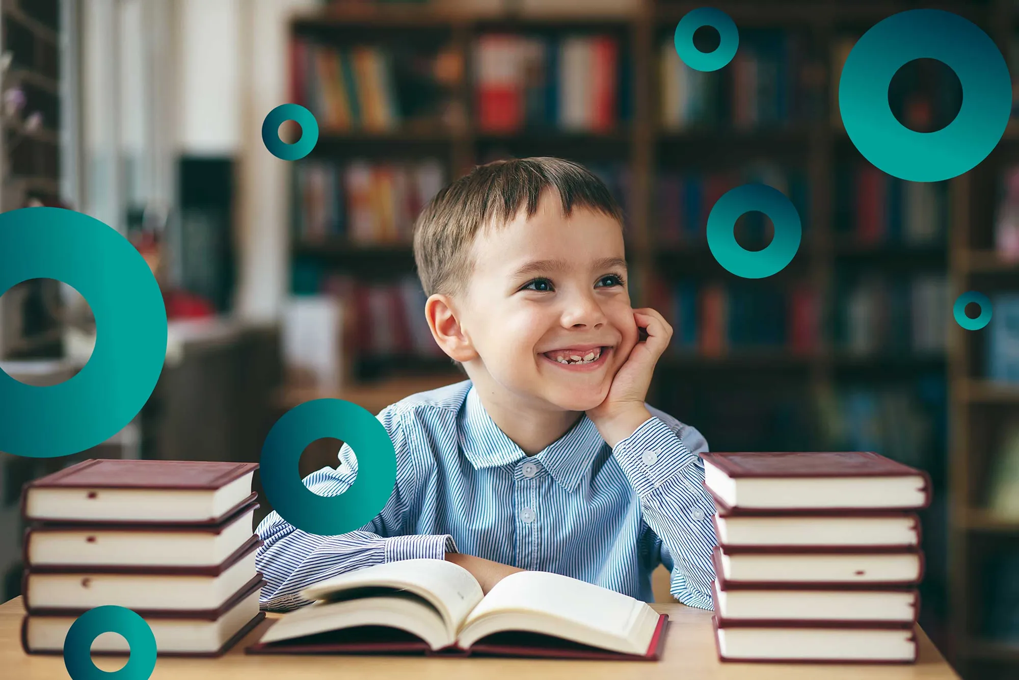 Smiling young boy sitting at a table with an open book and stacks of books in a library.