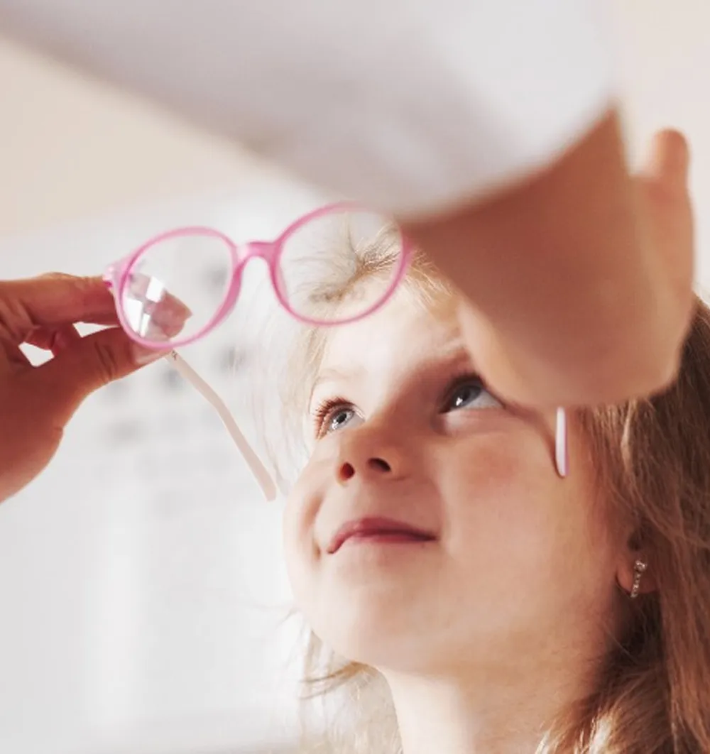 Child looking up attentively as an adult holds pink eyeglasses near their face.
