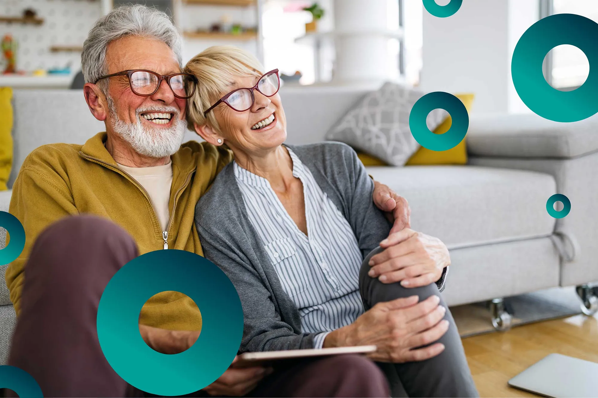 Smiling elderly couple sitting on floor, leaning close, holding tablet in a cozy living room.