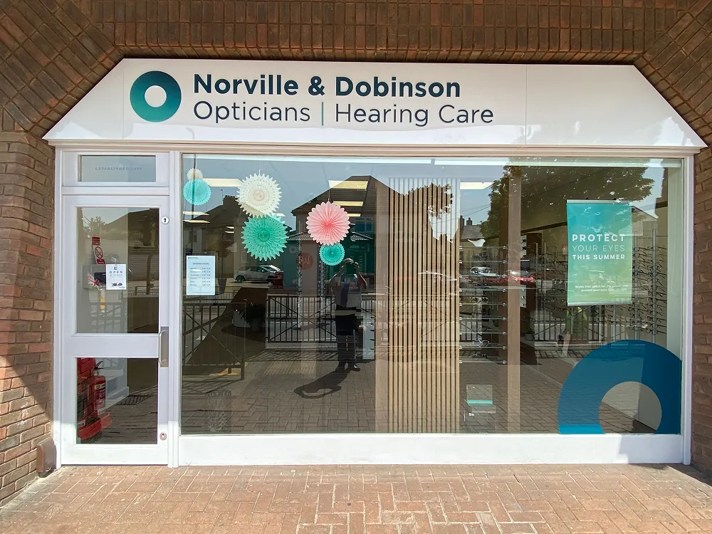 Front glass of Norville & Dobinson opticians and hearing care shop with promotional summer eye care poster.