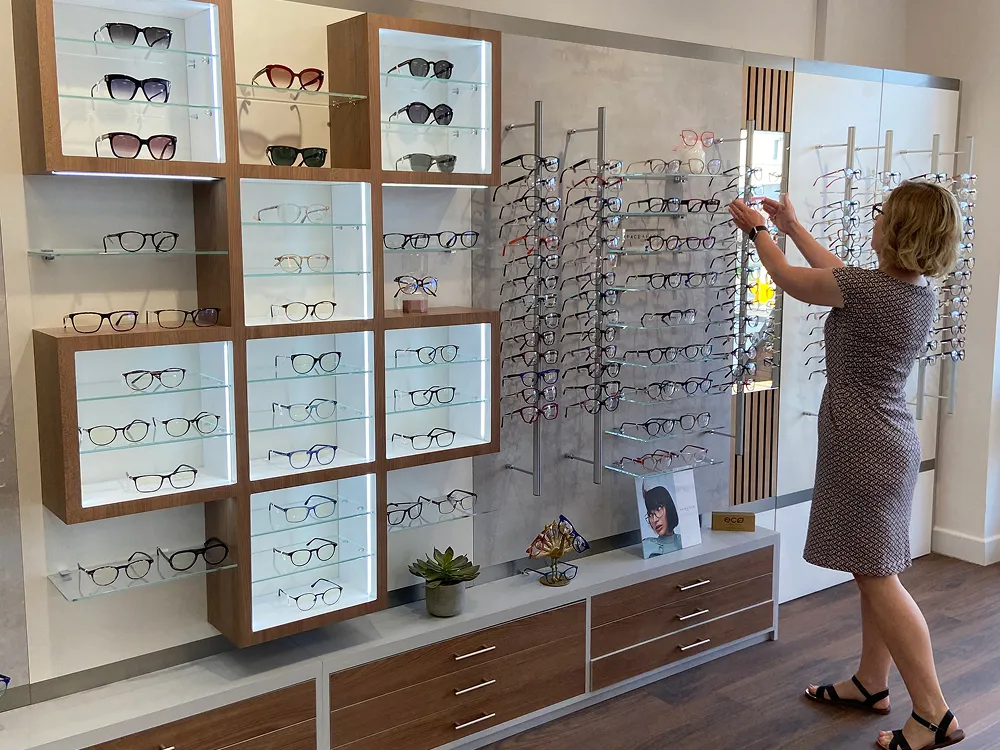 Woman in dress browsing multiple eyeglasses displayed on shelves and stands in optician store.