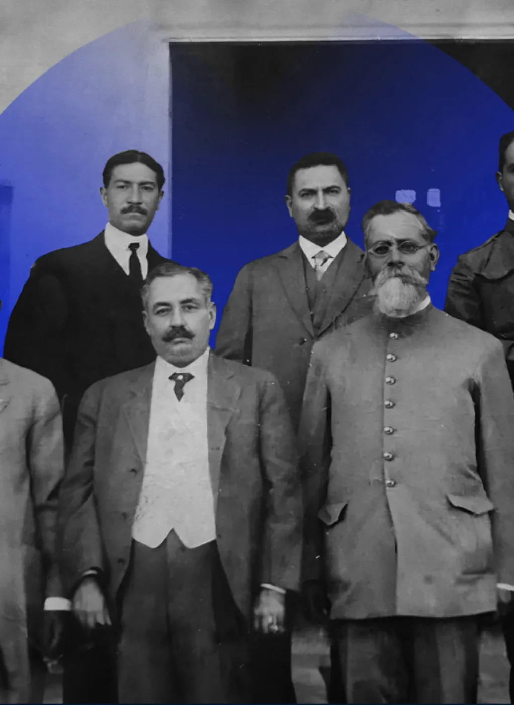 Black and white photo of five men in early 20th century formal attire posing in front of a building entrance.