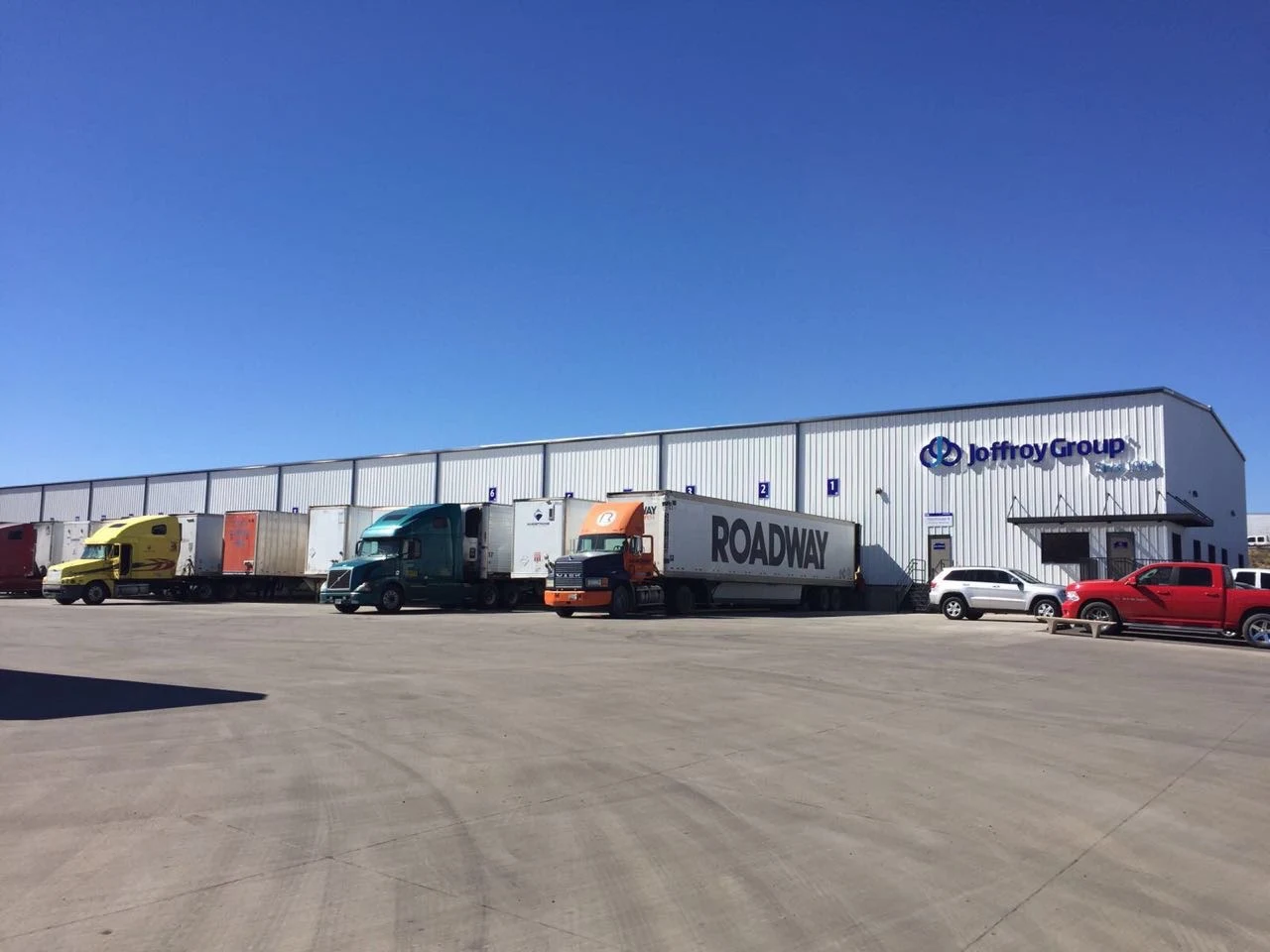Loading dock of Joffroy Group warehouse with trucks and cars parked under clear blue sky.