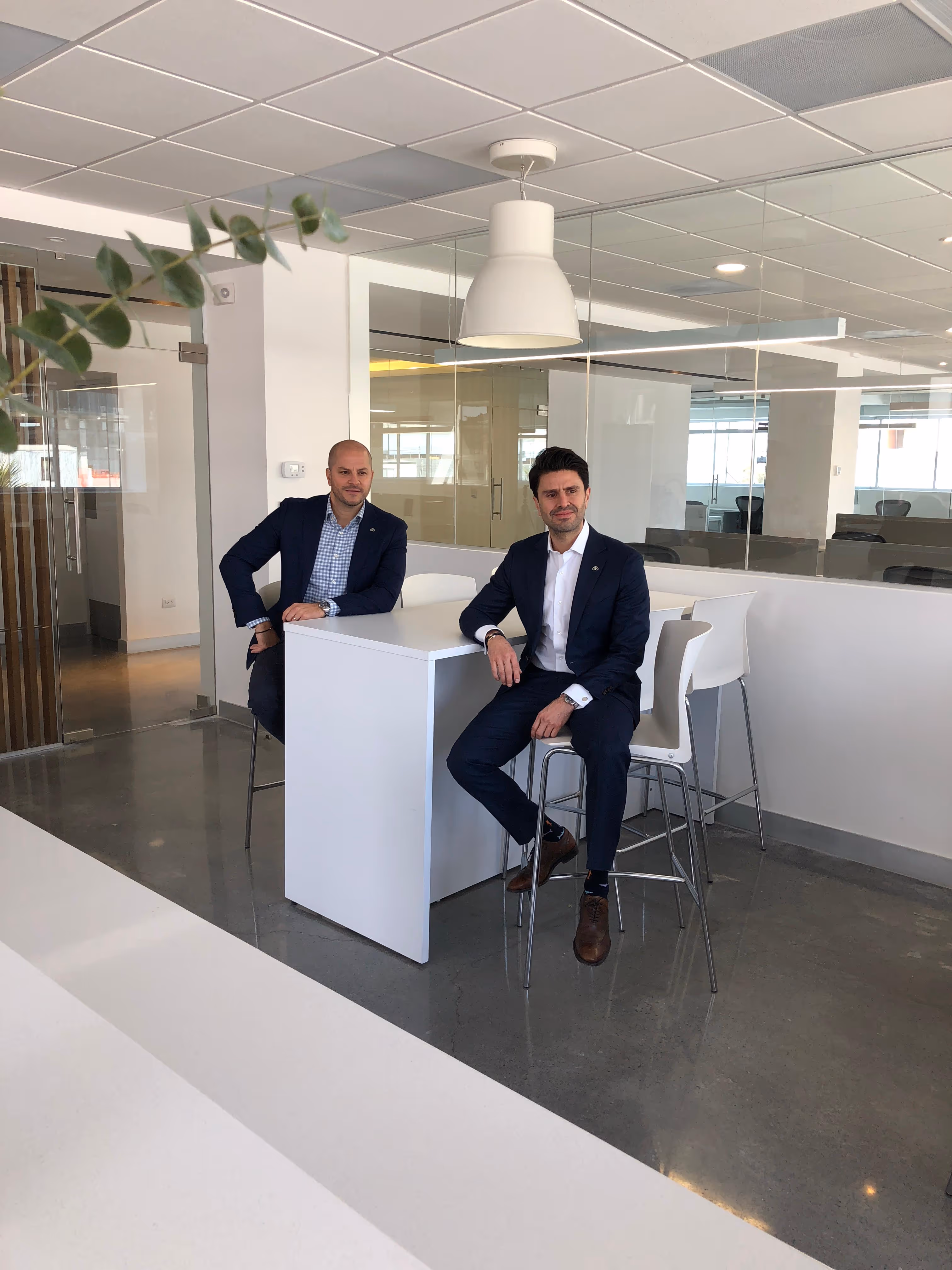Two men in suits sitting on stools at a white high table in a modern office space.