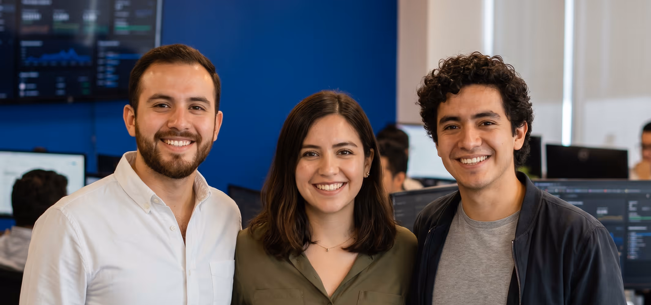 Three young adults smiling, standing close in an office with computer monitors behind them.