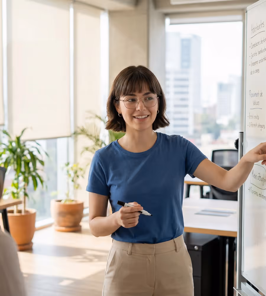 Young woman with glasses in blue shirt writing on whiteboard in bright office with plants.