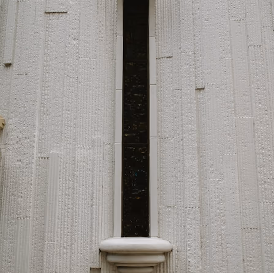 Narrow vertical window with dark glass set in a white textured wall with decorative molding at the base.