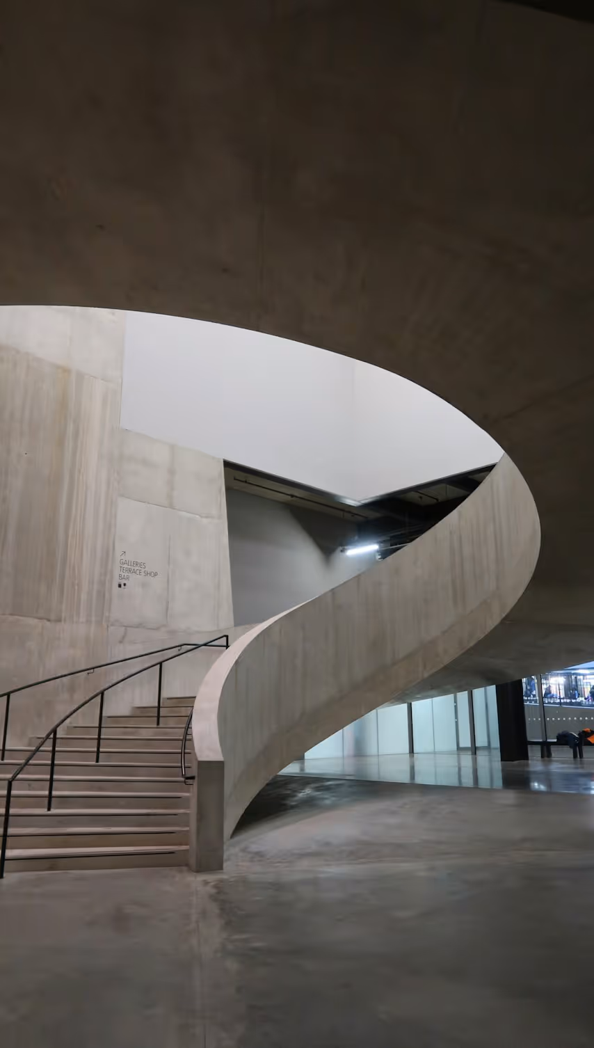 Modern concrete staircase with glass railing and large concrete beams overhead.