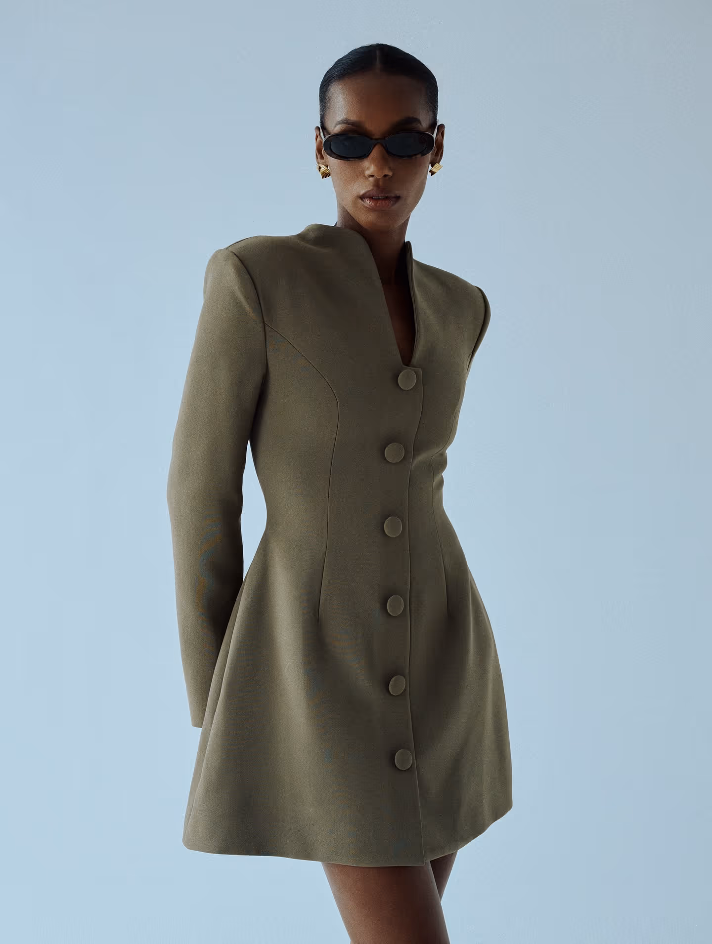 Woman wearing an olive green fitted coat dress with large buttons, black sunglasses, and gold earrings against a light blue background.