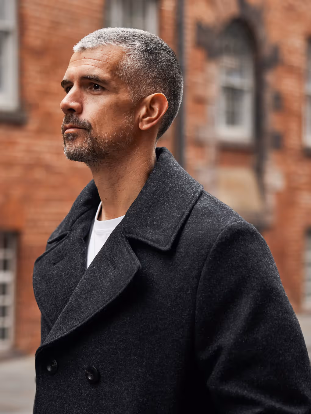 Serious middle-aged man with short gray hair and beard wearing a dark coat in front of a brick building.