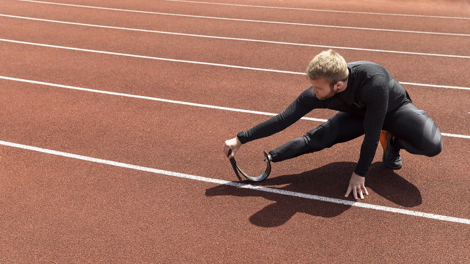 Male athlete with a prosthetic running blade stretching on a red outdoor track.