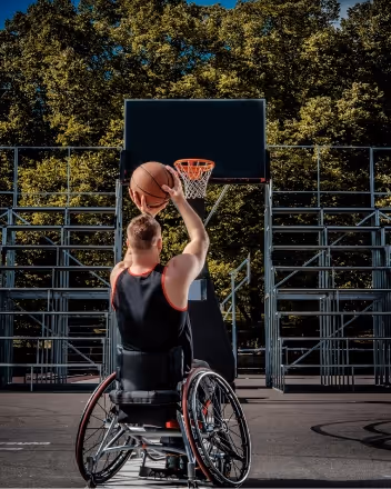 Man in wheelchair shooting a basketball toward an outdoor hoop with trees in the background.