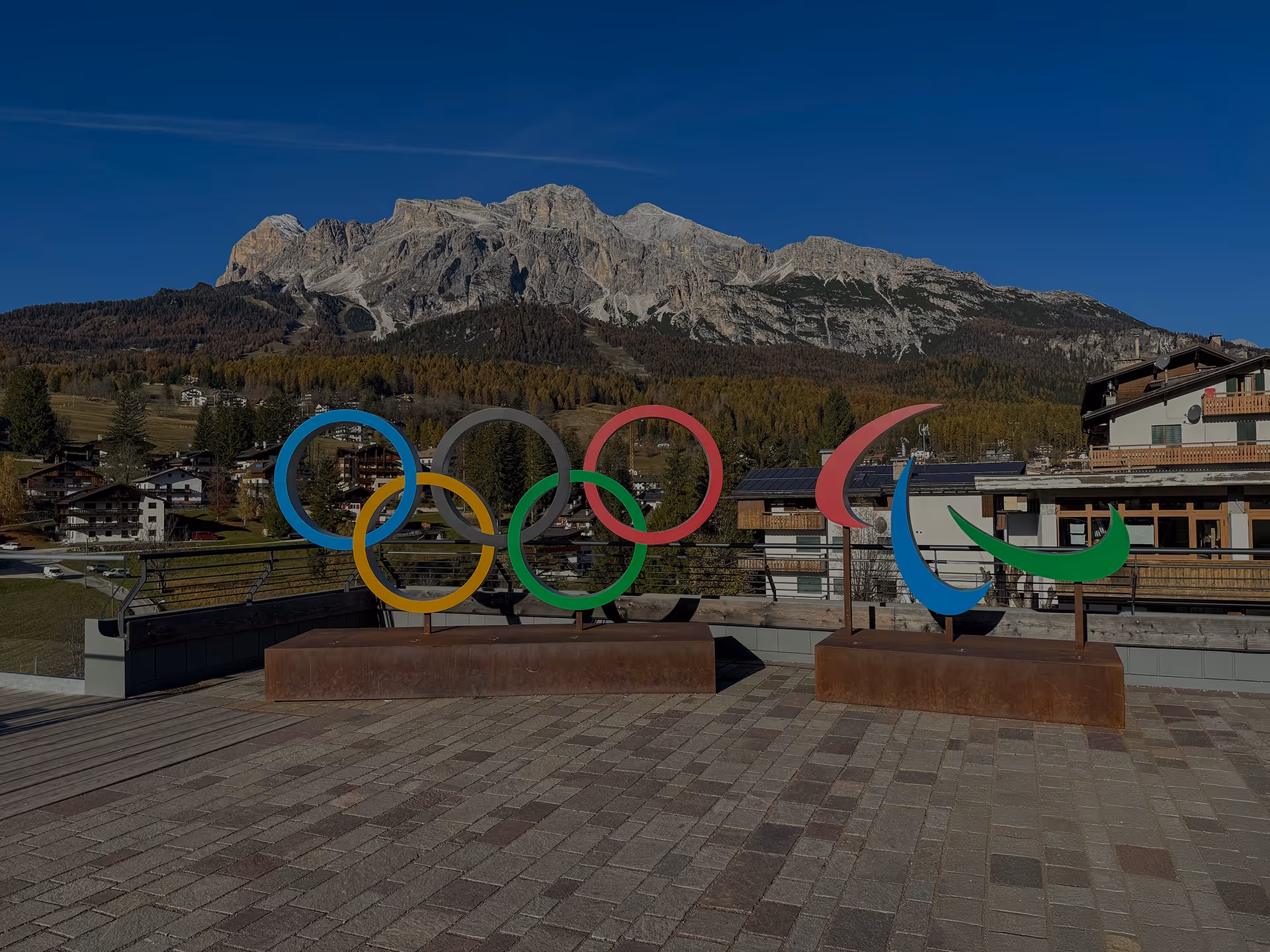Sculptures of the Olympic rings and Paralympic symbol displayed outdoors with mountains and alpine buildings in the background.