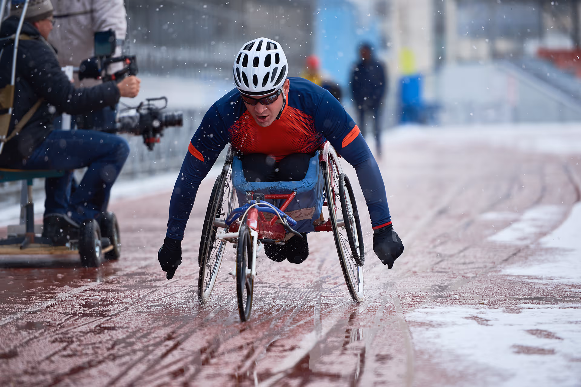 Athlete in a racing wheelchair competing on a wet track during snowy weather, wearing a white helmet and red and blue sportswear.