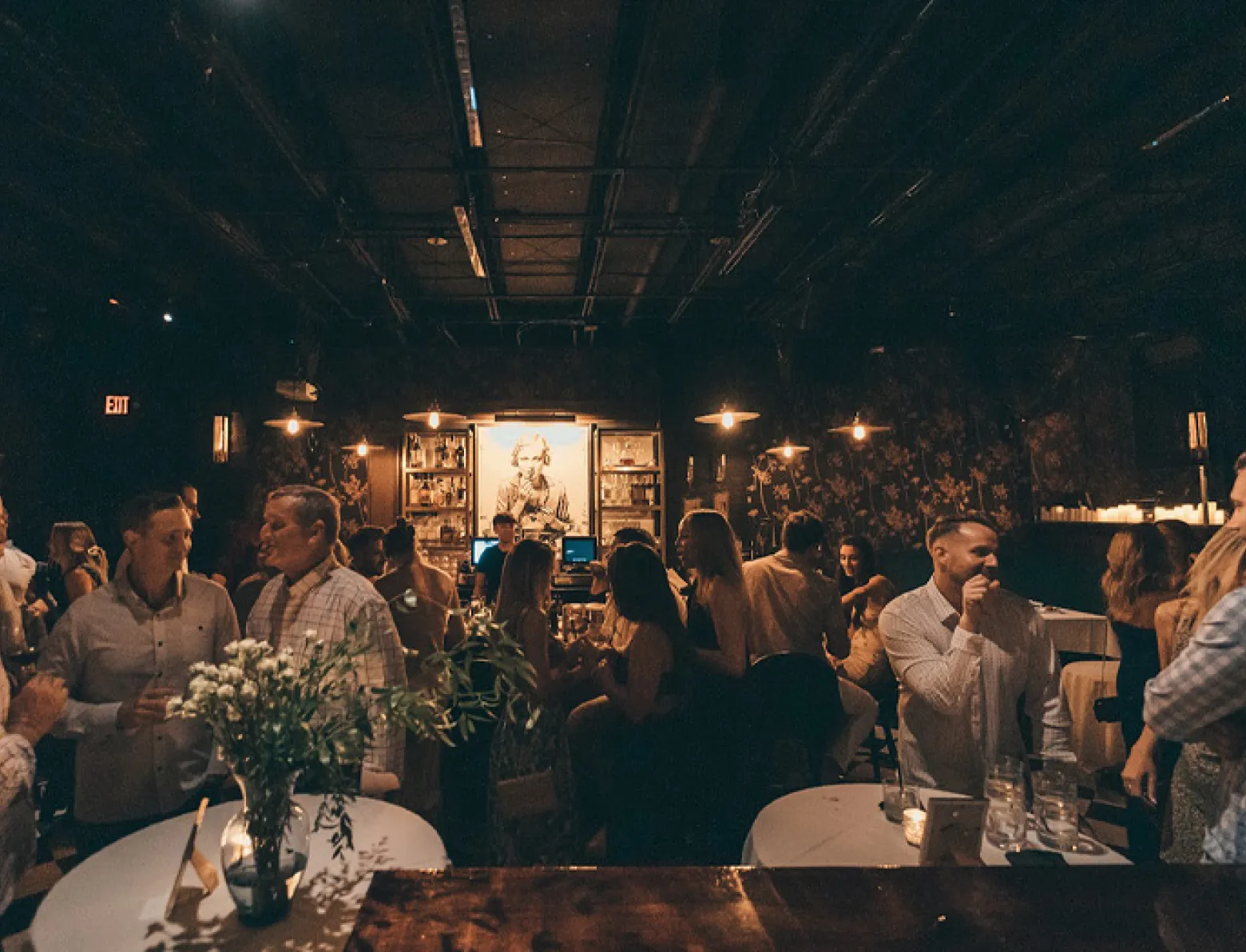 Dimly lit bar with people socializing around tables, warm hanging lights, and floral wallpaper.
