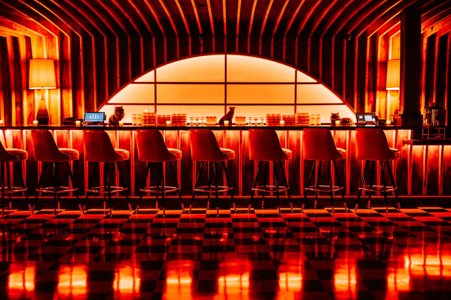 Red-lit bar interior with high stools, wooden paneled walls, checkerboard floor, and lamps on either side.