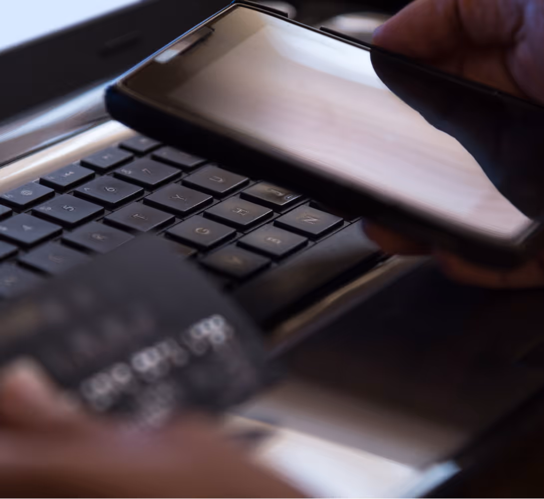 Close-up of a person holding a smartphone over a laptop keyboard with a blurred credit card in hand.
