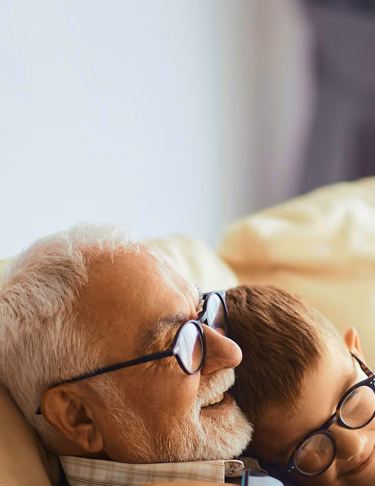 Elderly man and young boy wearing glasses, smiling and leaning close together on a couch.