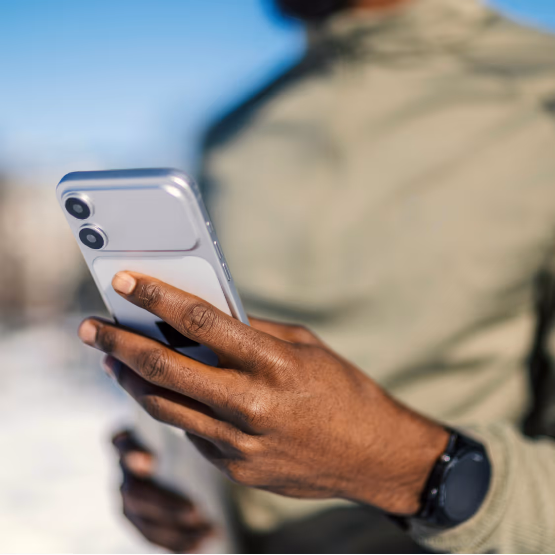 Close-up of a hand holding a silver smartphone outdoors with a blurred person in a green jacket in the background.