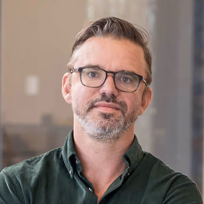 Man with short brown and gray hair, wearing black glasses and a dark green button-up shirt, posing indoors against a blurred background.