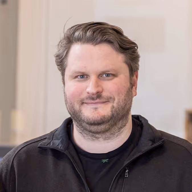 Smiling man with short wavy hair and beard wearing a black zip-up jacket.