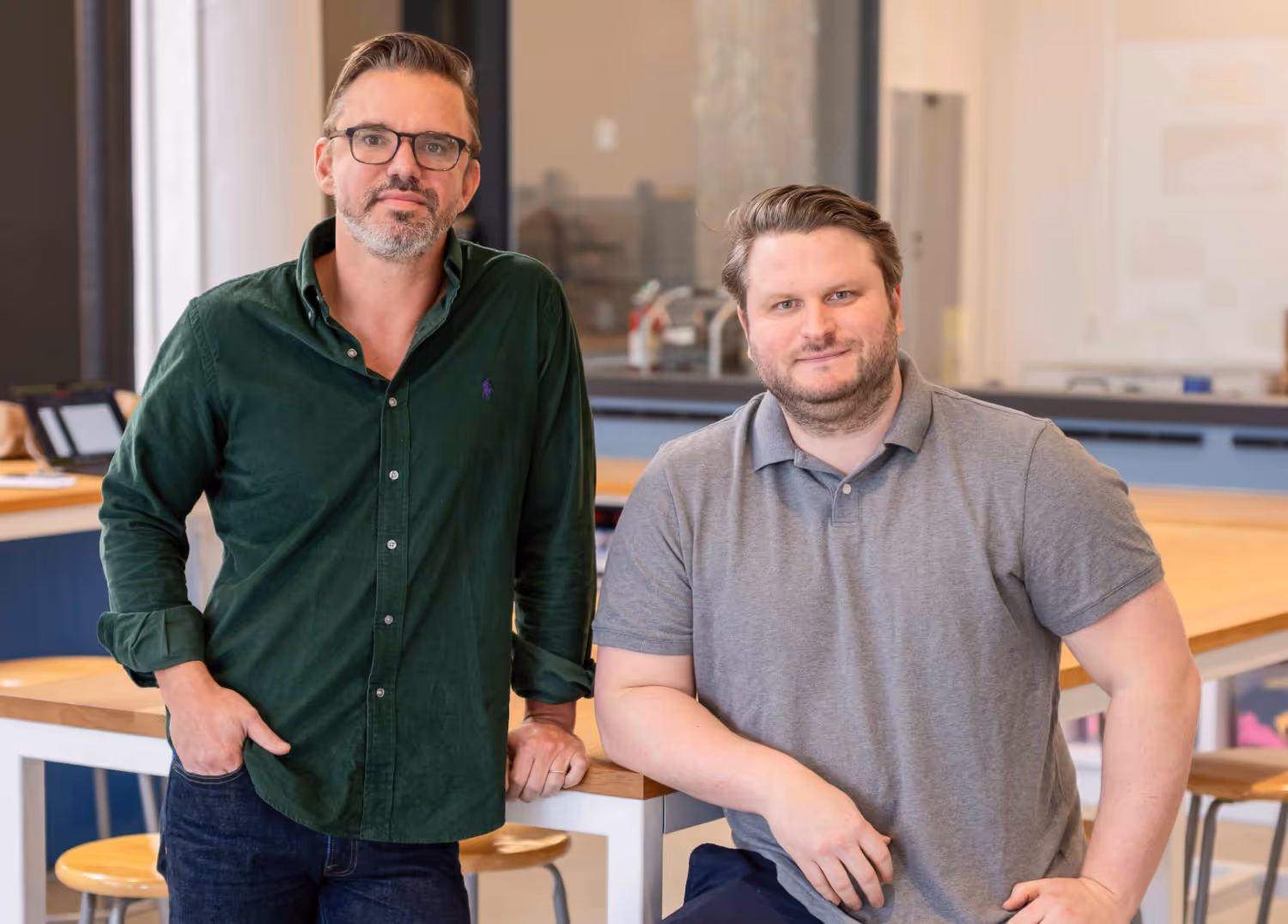 Two men posing casually in an office setting, one standing with hands in pockets wearing a green shirt and glasses, the other sitting wearing a gray polo shirt.